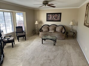 Una sala de estar con un sofá beige, dos sillones, una mesa de centro de vidrio y dos mesas auxiliares con lámparas.