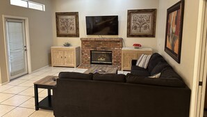 A living room with a brick fireplace, a black sofa, a TV mounted above the fireplace, and decorative wall art.