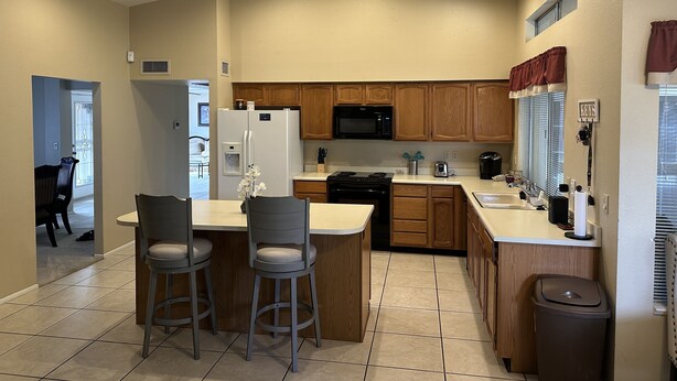 A modern kitchen with wooden cabinets, a refrigerator, a microwave, and a sink. There are two bar stools and a small island with a vase of flowers.