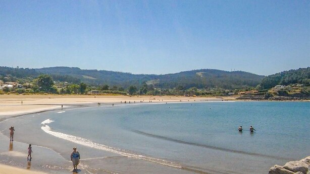 Ein Strand mit blauem Himmel, sanften Wellen und einer hügeligen Landschaft im Hintergrund.