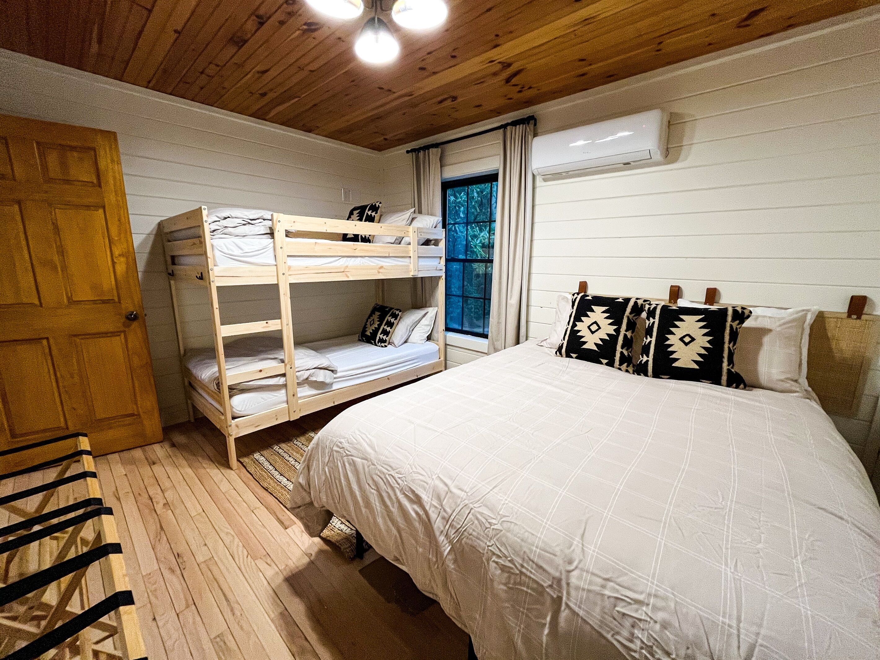 Un dormitorio con literas, una cama grande, un ventanal con cortinas y un equipo de aire acondicionado.