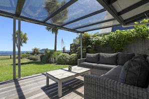 A covered outdoor seating area with a view of the ocean, a wooden table, and a wicker sofa. - Hannah's Bay Lakefront - Kayaks, Sleeps 8 (Rotorua)