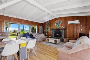 A spacious living room with a fireplace, wooden walls, and a large window overlooking a garden. - Hannah's Bay Lakefront - Kayaks, Sleeps 8 (Rotorua)