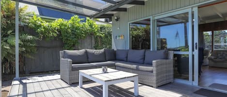 An outdoor patio with a white table and grey sofa, surrounded by a wooden deck and a glass enclosure.