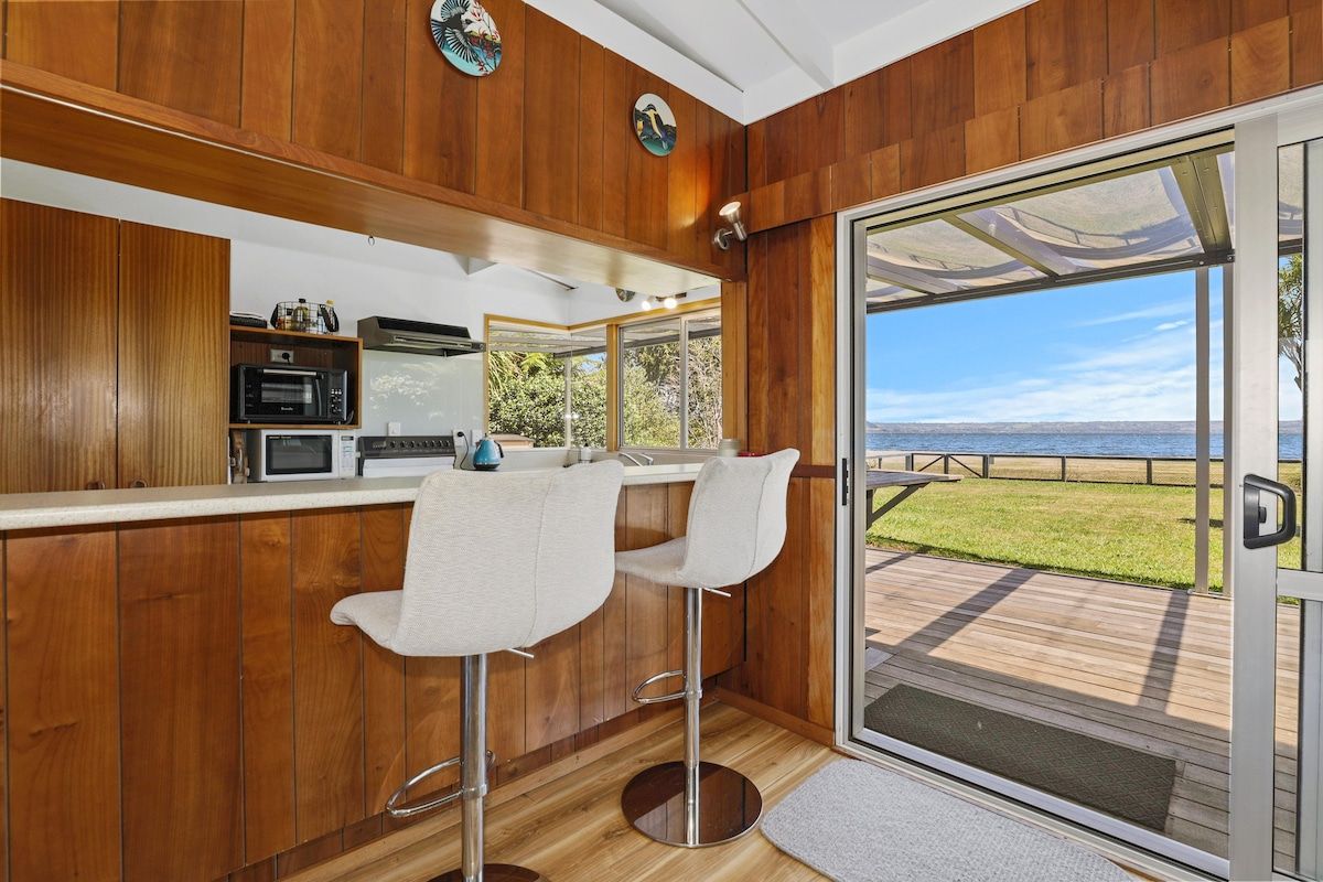 A kitchen with wooden paneling, a bar area with two stools, and a view of the sea through a glass door.