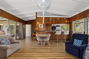 A modern interior with a wooden floor, a dining area with a table and chairs, a sofa, a blue armchair, and a kitchen area with a refrigerator and stove. - Hannah's Bay Lakefront - Kayaks, Sleeps 8 (Rotorua)
