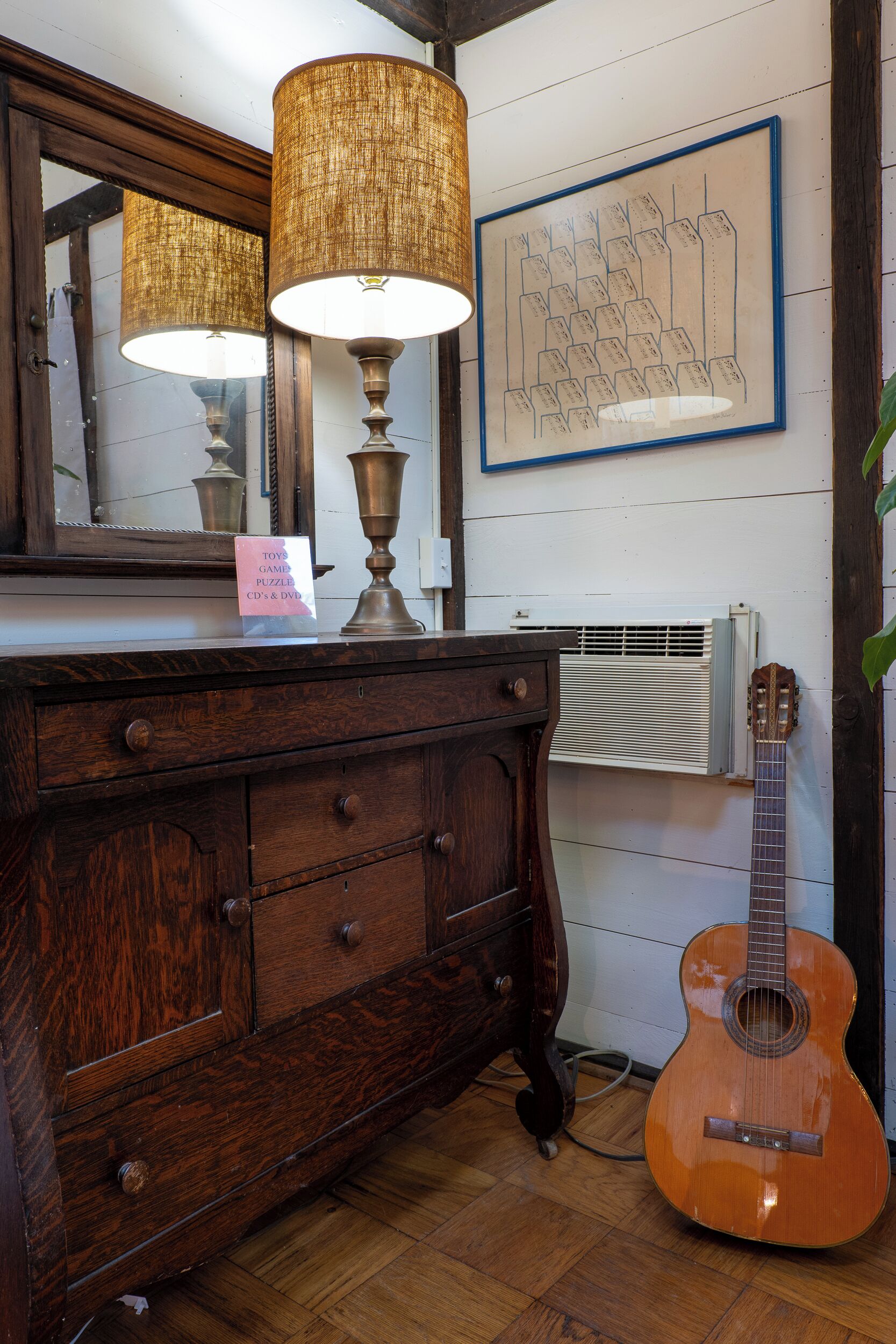 A wooden dresser with a mirror, a lamp, a guitar, and a framed picture on the wall.