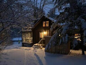 Una cabaña de madera con un porche con paredes de vidrio, rodeada de árboles cubiertos de nieve.