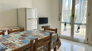 A dining area with a table covered in a patterned tablecloth, wooden chairs, a white refrigerator, a television on a small stand, and a sliding glass door with sheer curtains. - F002 - Fano, three-room apartment with porch 30m from the sea (Fano)