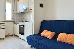 Eine kleine Küche mit Herd, Backofen und Spüle, ein blaues Sofa mit orangefarbenen Kissen und ein Fenster mit Jalousien.