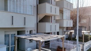 Un edificio residencial de varios pisos con balcones, un espacio al aire libre techado para sentarse y una cerca blanca de piquetes.