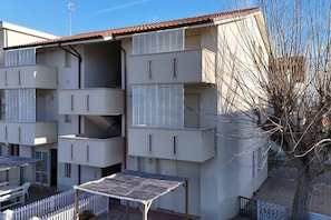 A multi-story residential building with balconies, a covered outdoor seating area, and a tree in the foreground. - F005 - Fano, three-room apartment with small terrace 30m from the sea (Fano)