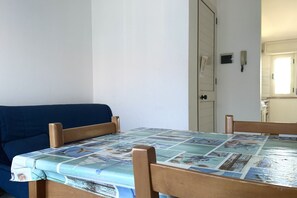 Een kamer met een houten tafel, een blauwe bank en een deur die naar een keuken leidt.