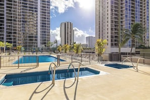 Un espace piscine aménagé sur le toit, comprenant plusieurs piscines, entouré de grands immeubles et de palmiers.