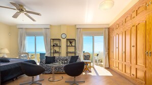 Een kamer met een bed, twee stoelen, een bank, een plafondventilator, een klok en uitzicht op zee.