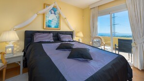 Een slaapkamer met een bed, nachtkastje, stoel, raam met gordijnen en balkon met een stoel.