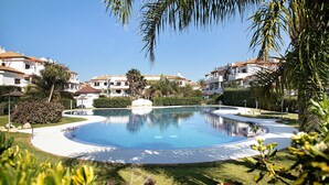 A swimming pool surrounded by a well-maintained lawn, palm trees, and residential buildings.