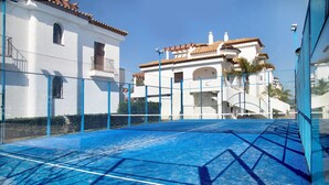 A tennis court with a net, surrounded by a blue fence, and buildings with white walls and red-tiled roofs in the background.