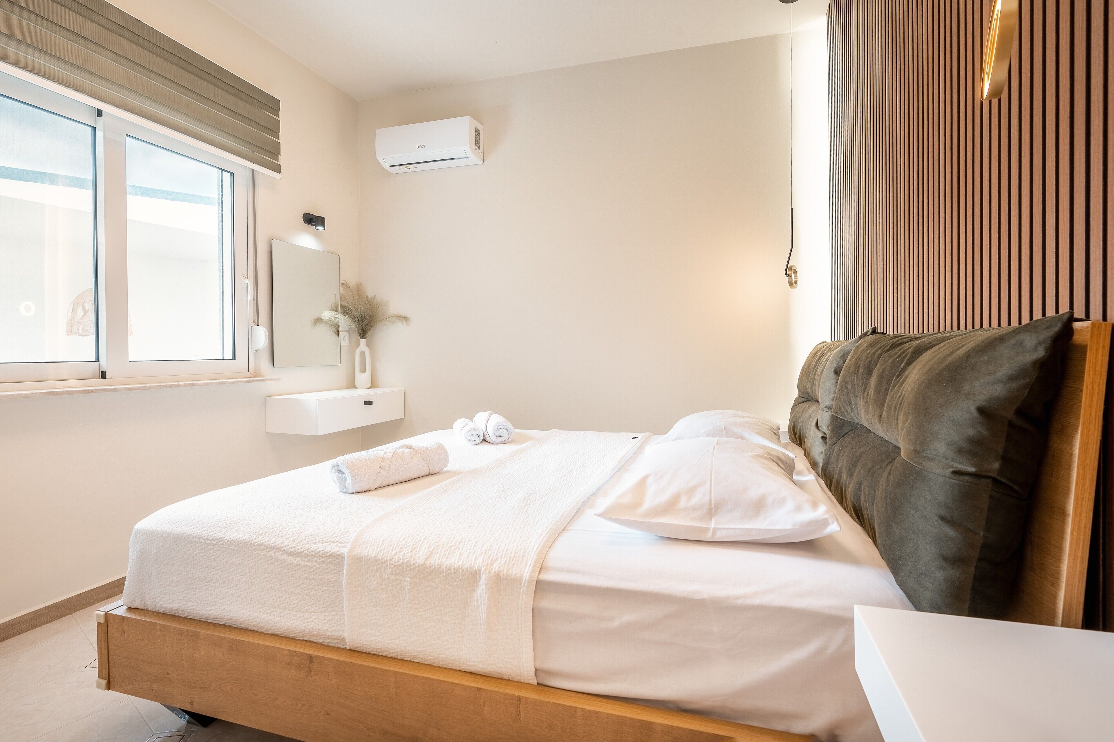 Ein ordentlich bezogenes Bett mit weißen Leinen, ein Nachttisch, eine Wand-Klimaanlage, ein Fenster mit Jalousien und ein Holzkopfschild.