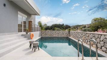 A modern outdoor pool area with stone walls, steps leading to a building, and a clear view of the surrounding landscape.