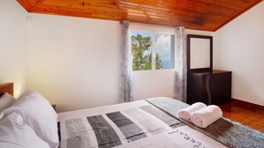 Ein Schlafzimmer mit Holzbalkendecke, einem Bett mit Decke, einem Fenster mit Vorhängen, einem Schminktisch und einem Spiegel.