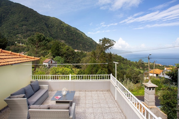 A balcony with a sofa, table, and railing overlooking a mountainous landscape.