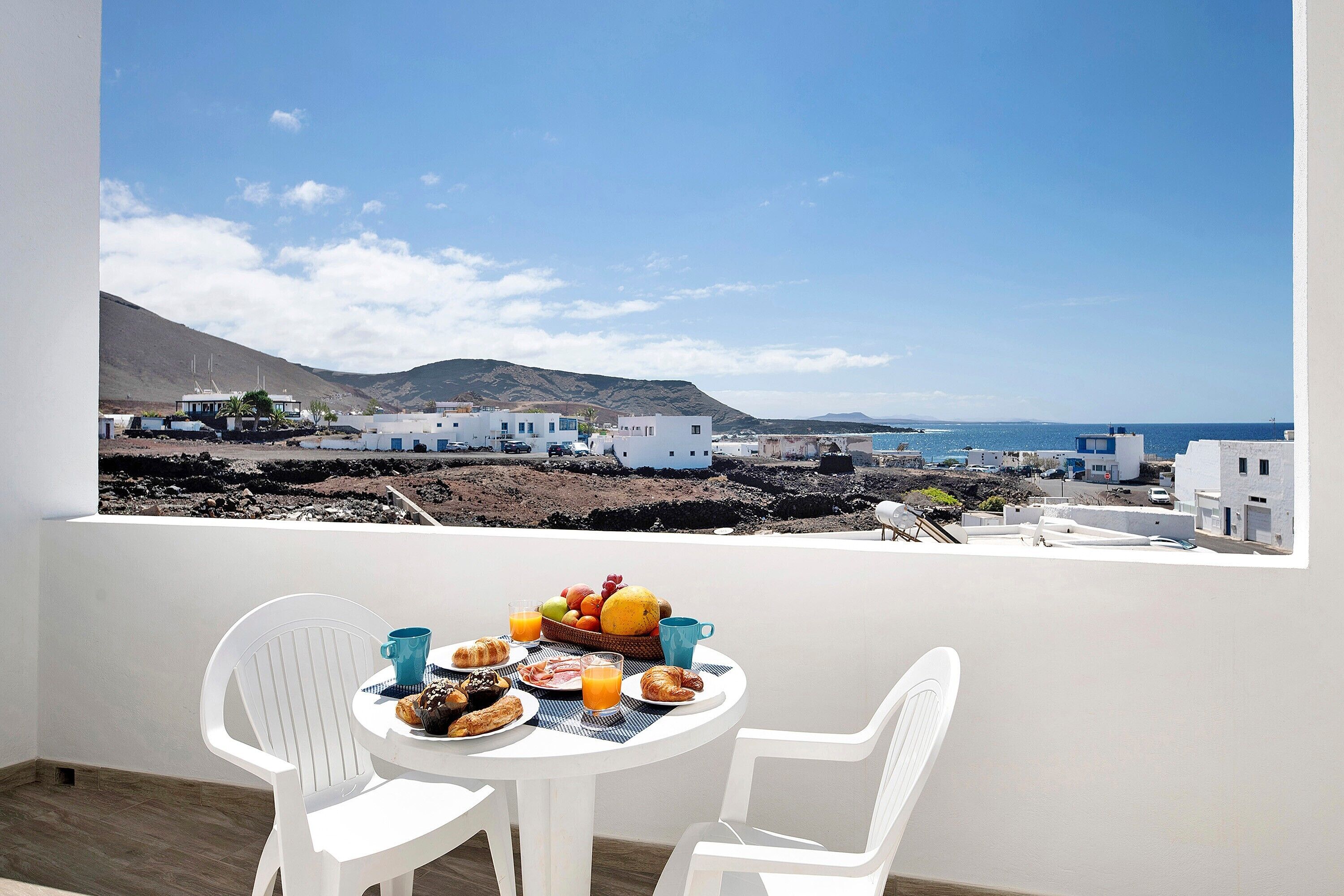 A balcony with a table set for breakfast, overlooking a coastal village with white buildings and a clear sky.