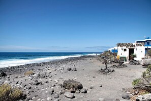 Uma praia com costa rochosa, um edifício branco com detalhes azuis e um céu azul límpido.