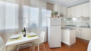 A compact kitchen with a refrigerator, a dining table with chairs, a small island, and a window with sheer curtains.