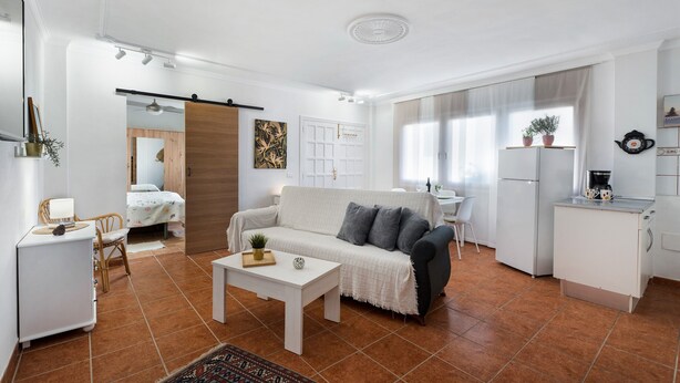 A modern living room with a sofa, coffee table, and a kitchen area with a refrigerator and sink.