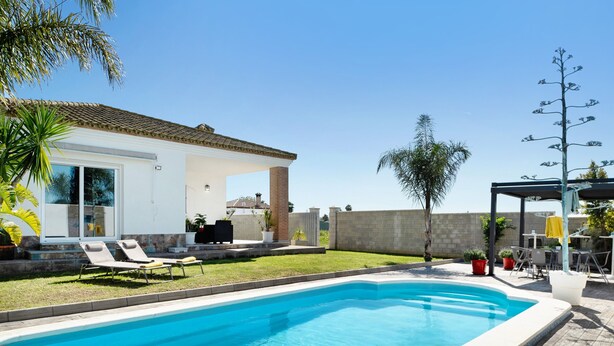 A swimming pool with lounge chairs, a covered patio, and a house with a tiled roof.