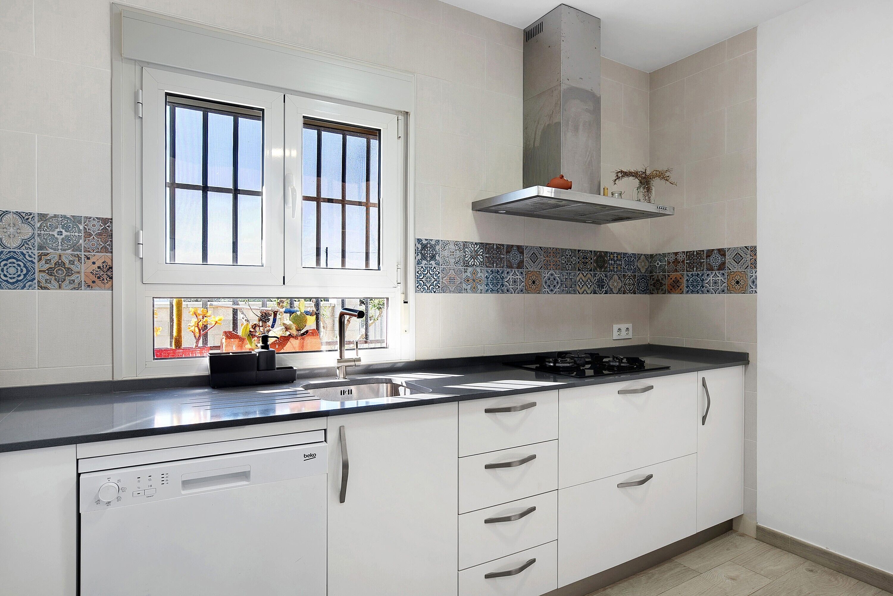 Una cocina con armarios blancos, encimera negra, ventana con persianas y salpicadero de azulejos.