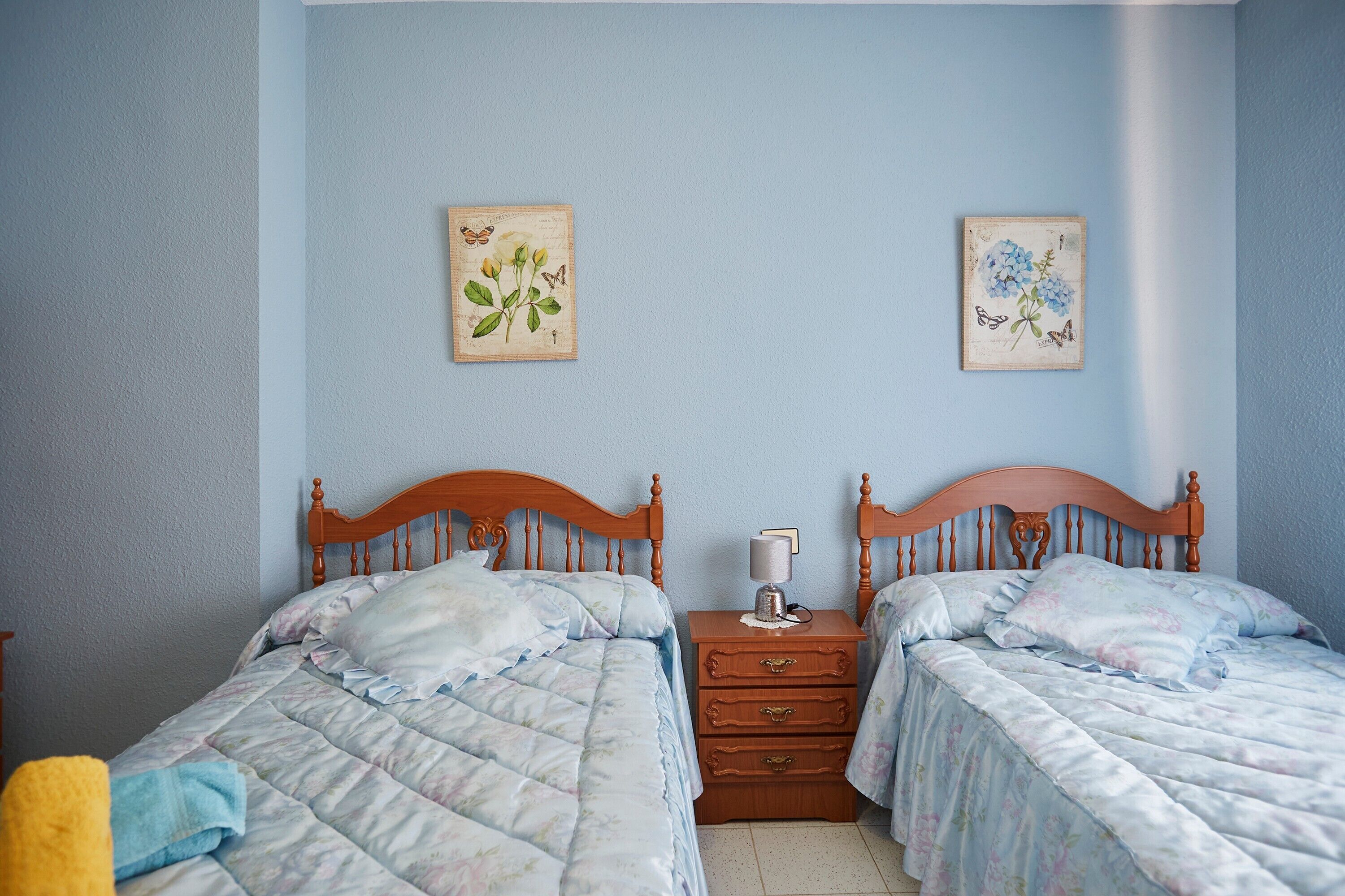 Un dormitorio con dos camas de madera, una mesita de noche con lámpara y decoración floral en la pared.