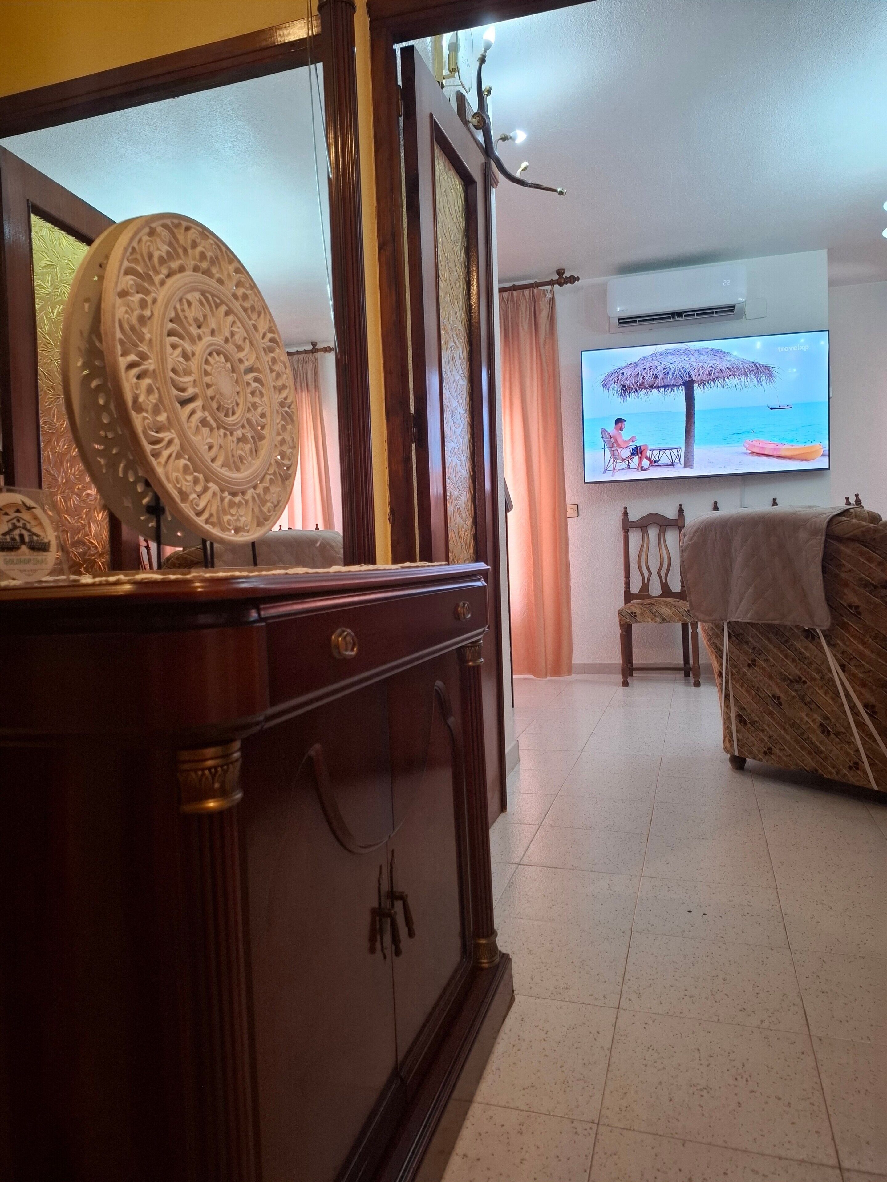 Una habitación con un armario de madera, un televisor que muestra una escena de playa, una silla y una cama con una manta.