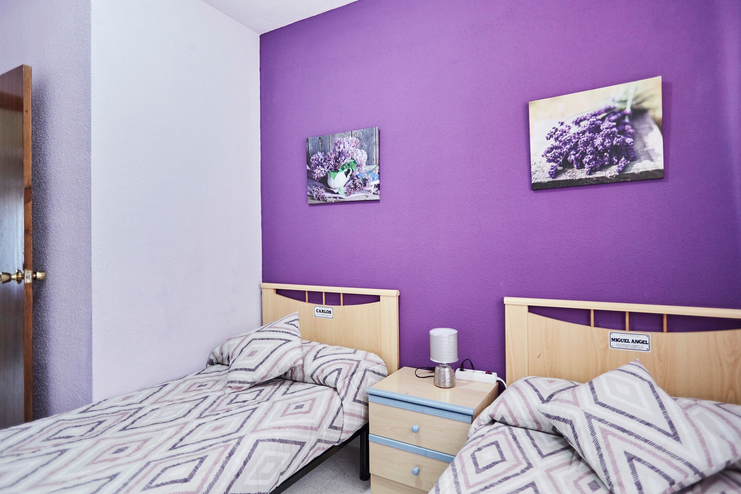 A bedroom with two single beds, a nightstand, a lamp, and two wall-mounted pictures.