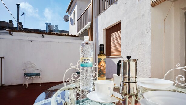 Une table dressée avec des assiettes, une tasse, une bouteille et une cafetière, sur un balcon. En arrière-plan, on aperçoit une chaise blanche et une chaise en métal.
