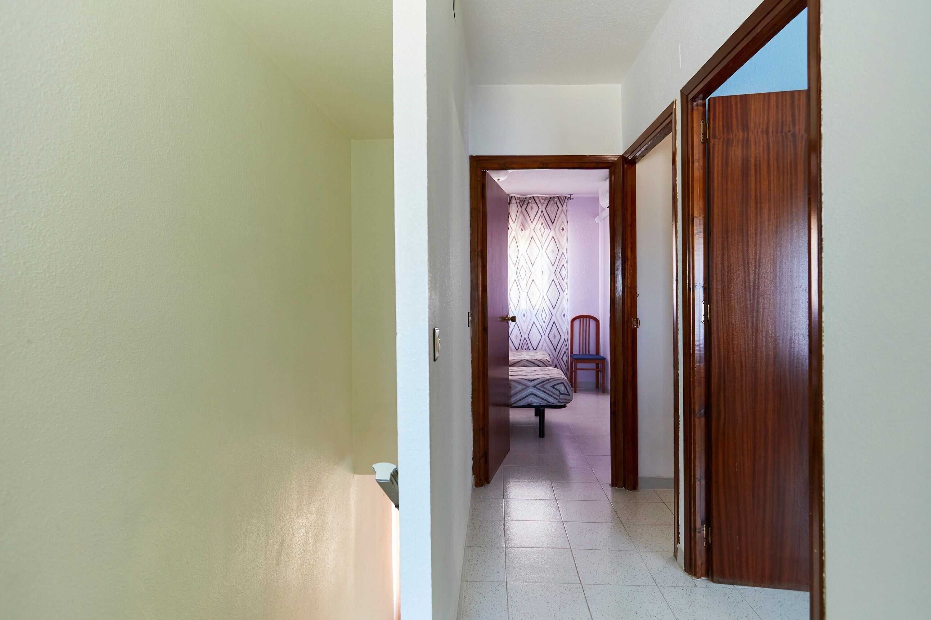 Un estrecho pasillo que conduce a un dormitorio con cama y sillón, puertas de madera y suelo de baldosas.