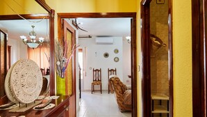 Un couloir avec une commode en bois, un miroir, un lustre et un escalier menant à une pièce où se trouvent un lit et des chaises.