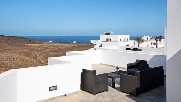 Eine Dachterrasse mit Gartenmöbeln, Blick auf das Meer und weiße Gebäude im Hintergrund.