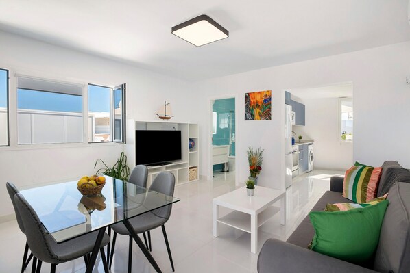 A modern living room with a glass dining table, grey sofa, and a flat-screen TV.