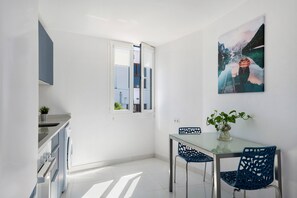 A modern kitchen with a glass dining table, blue chairs, and a painting of a mountain landscape.