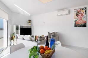 Una habitación moderna con un sofá blanco, una mesa de comedor con fruta y una botella, un televisor y un cuadro en la pared.