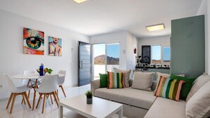 Una sala de estar moderna con un sofá, una mesa de comedor y sillas. Hay dos cuadros en la pared, una puerta que da al exterior y un área de cocina visible a través de una puerta abierta.