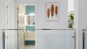 Un couloir d’hôtel moderne avec une rambarde en verre, une salle de bain aux accents turquoise et une œuvre d’art représentant une feuille, encadrée et accrochée au mur.