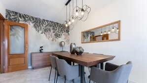 Een eetgedeelte met een houten tafel, grijs gestoffeerde stoelen, een decoratieve lamp, een spiegel en een behang met een patroon.