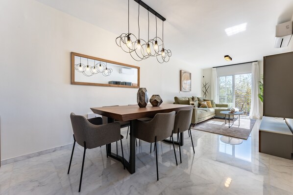 A modern dining area with a wooden table and chairs, a chandelier, a large mirror, and a sofa in the background.