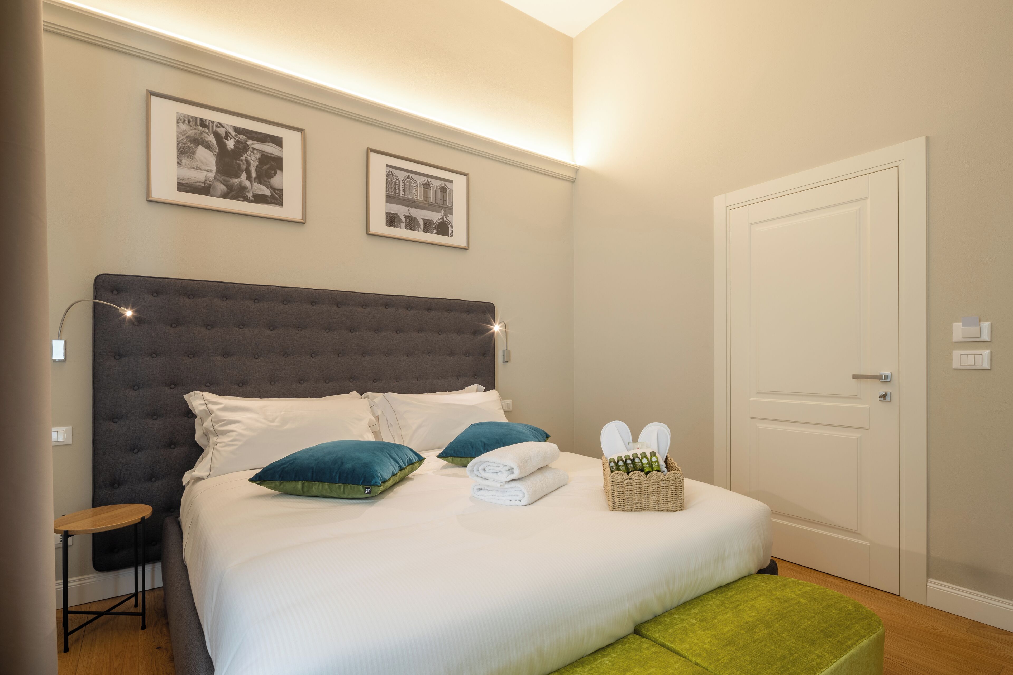 Una cama bien tendida con almohadas, una mesita de noche, una cesta con artículos de aseo y cuadros en la pared.