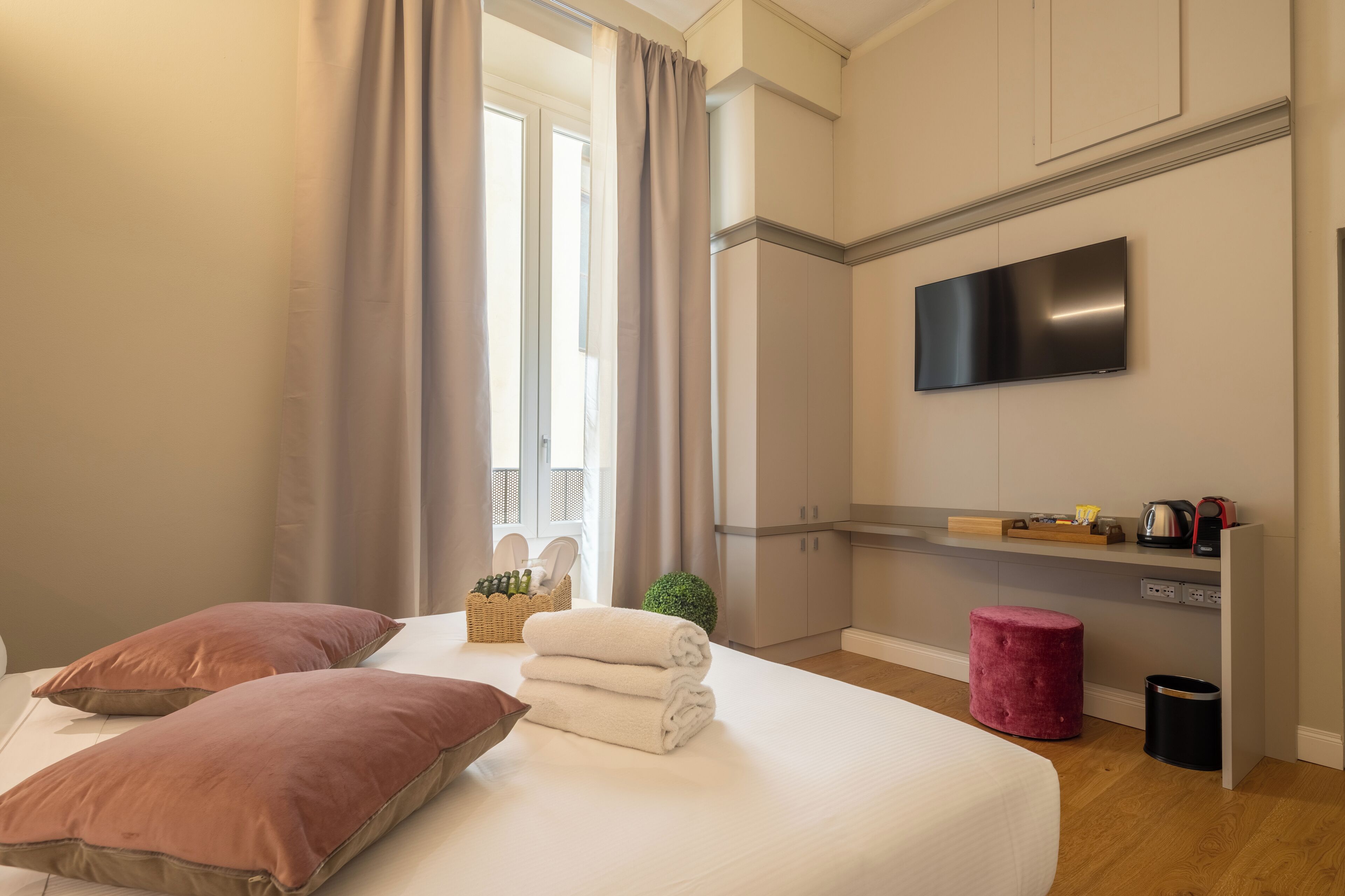 Quarto de hotel com cama, almofadas, televisão, uma pequena planta, um banquinho vermelho e um cesto de lixo.