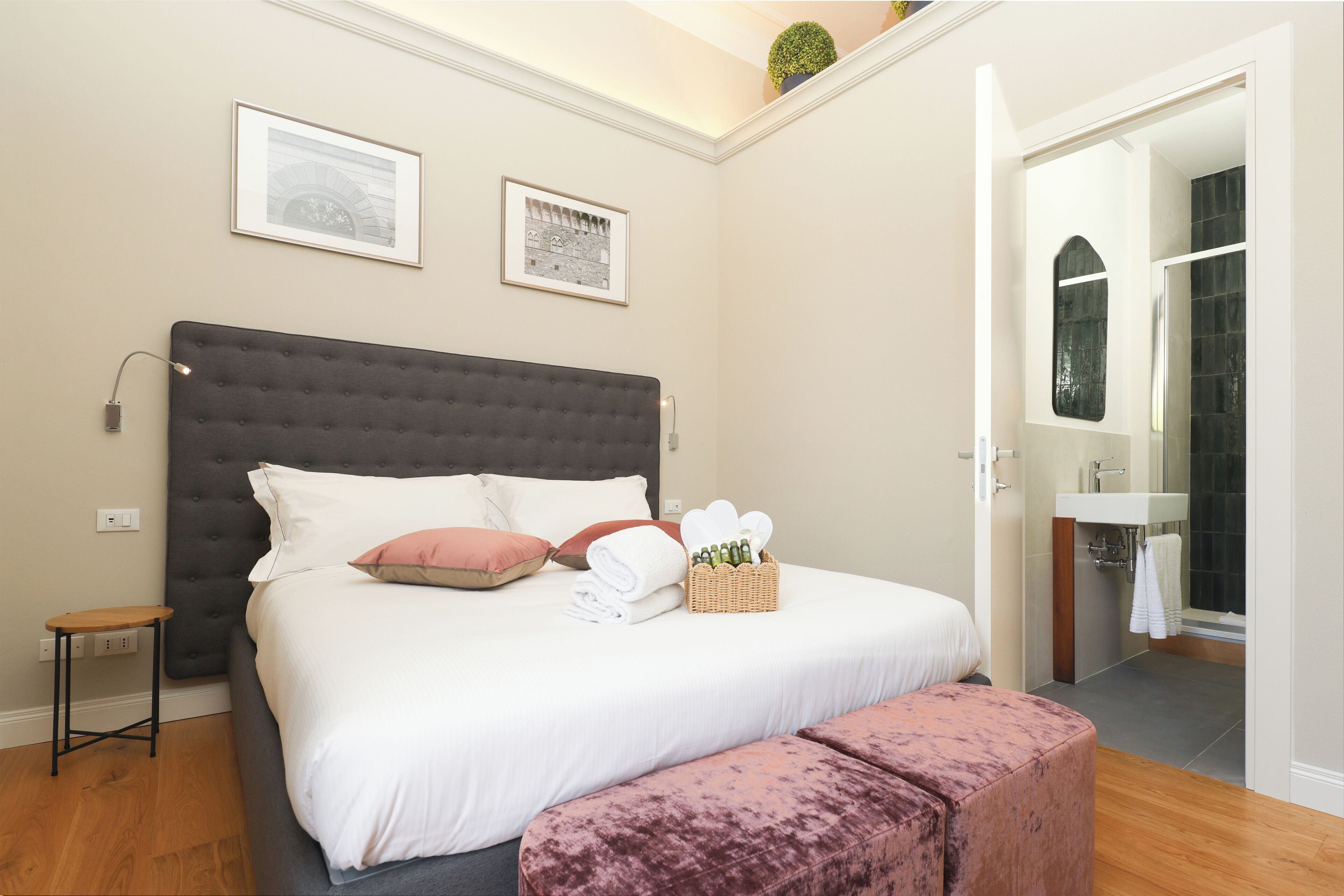 A bedroom with a large bed, a bedside table, a small stool, a bench, a basket of toiletries, and a bathroom visible through an open door.