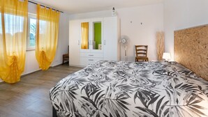 Quarto com cama, guarda-roupa, cadeira, ventilador e janela com cortinas.